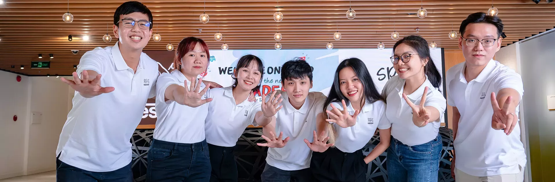 A group of six people in matching white shirts pose together, playfully making hand gestures against a bright