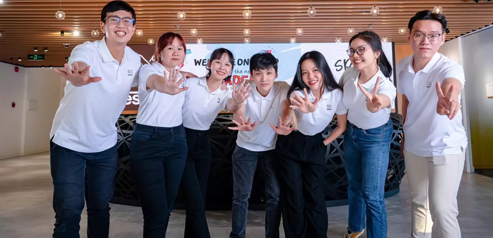 A group of six individuals wearing matching white polo shirts are playfully posing with their hands outstretched in a welcomi